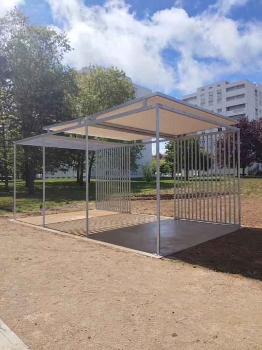 Abri-pergola en acier dont la toiture évoque un livre ouvert, sur une place