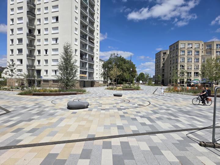 Lignes courbes en acier corten incrustées dans un dallage gris sur le parvis de la gare