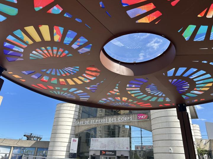 Sous-face d'une ombrière circulaire en acier découpé laissant passer la lumière à travers des motifs colorés, devant la gare de Saint-Pierre-des-Corps