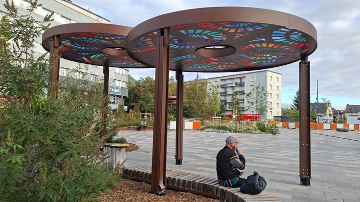 Voyageur assis sur un banc en lames de bois sous une ombrière circulaire en acier sur le parvis de la gare