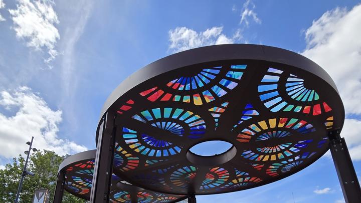 Ombrières circulaires en acier découpé aux motifs colorés vues d'en dessous sur un ciel bleu, parvis de la gare de Saint-Pierre-des-Corps