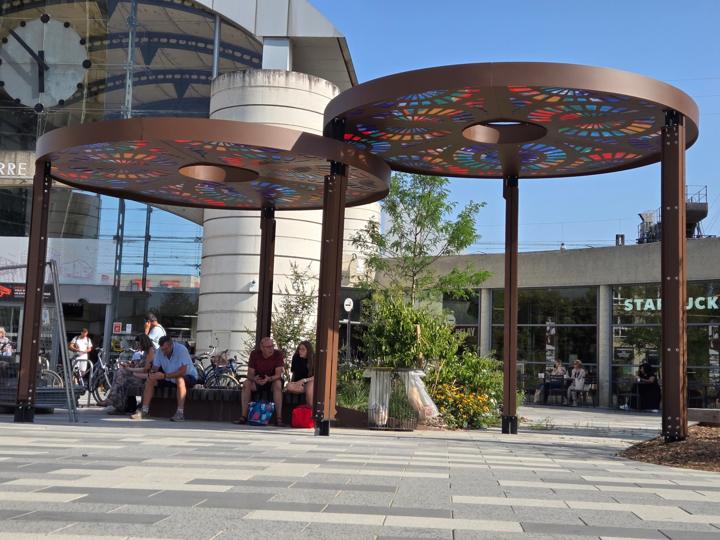 Deux ombrières circulaires en acier aux motifs colorés abritant des voyageurs assis sur des bancs devant la gare de Saint-Pierre-des-Corps