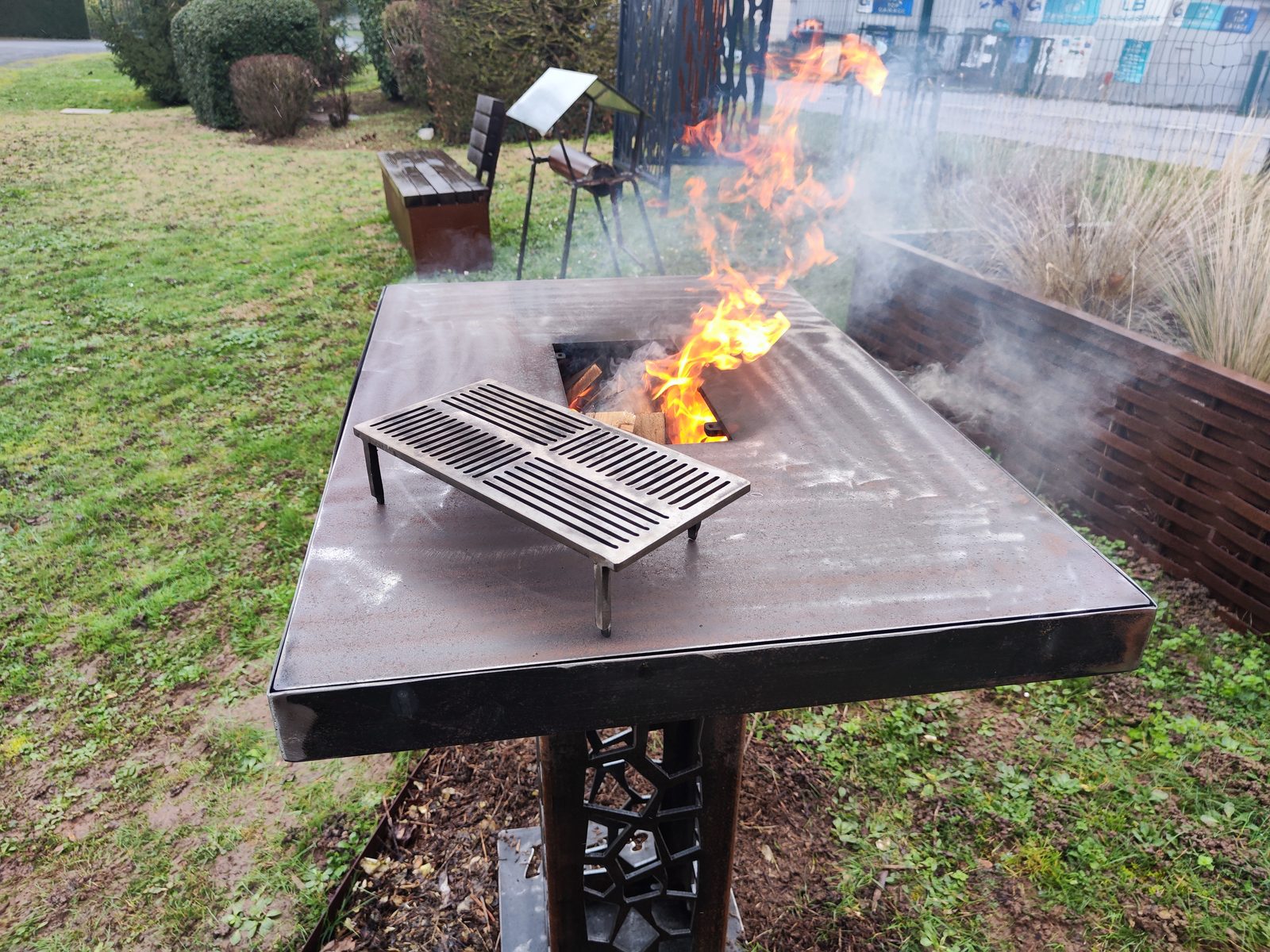 Table-brasero rectangulaire en acier avec foyer central allumé et grille de cuisson posée sur le plateau